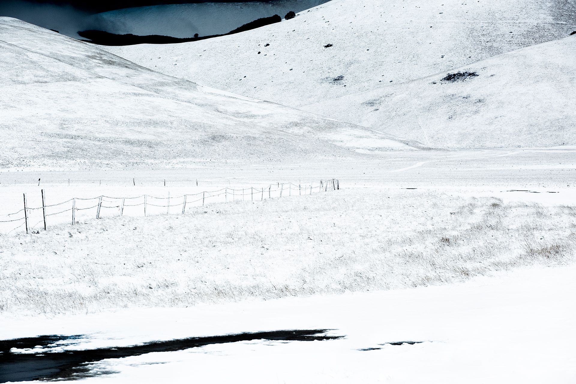 Staccionata e prato innevato sotto le colline, con una pozza d'acqua in primo piano